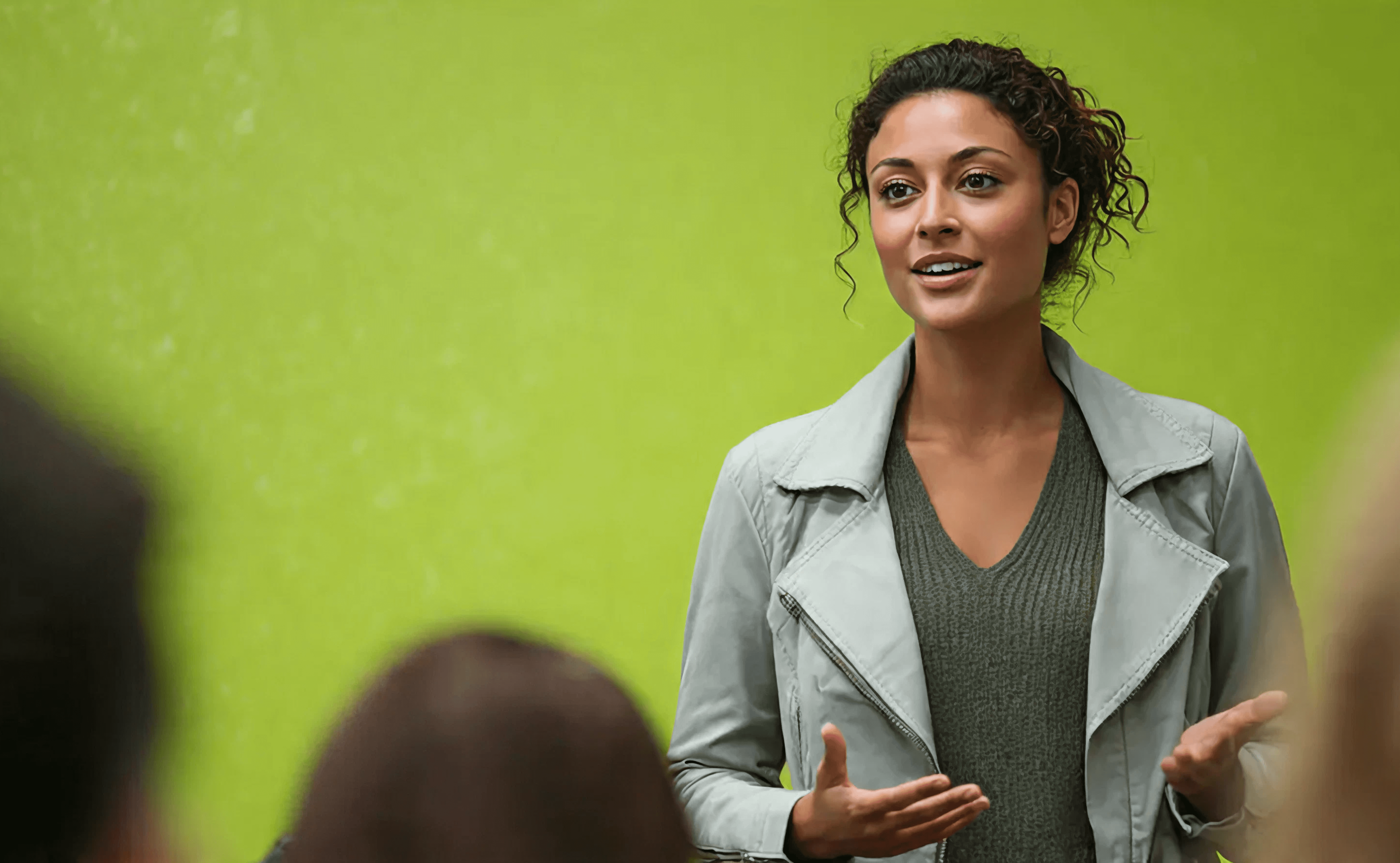 Woman speaker gesturing at the audience against a bright green wall background.
