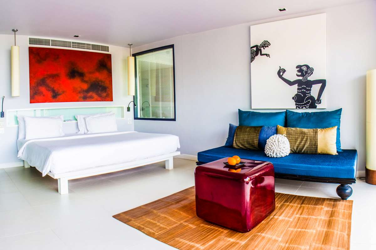 A modern hotel room features a white bed with red and orange abstract art above it and a blue sofa with colorful pillows.