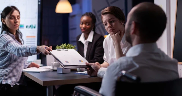 Pessoas em volta de uma mesa de reunião com laptop, uma delas entregando um documento