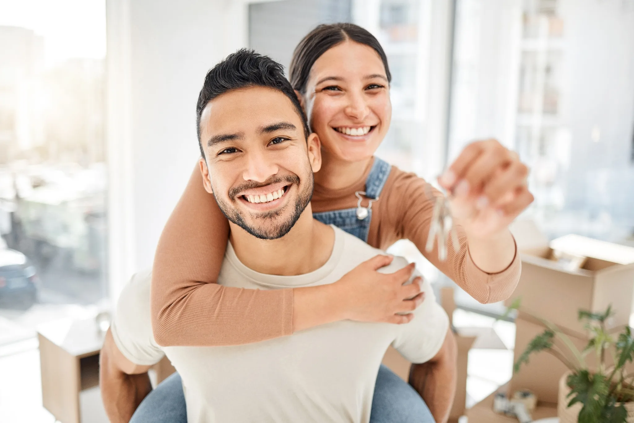 Smiling couple, woman piggy back riding at the back of the male, right hand hugging the male and left hand slightly raised forward holding a house key