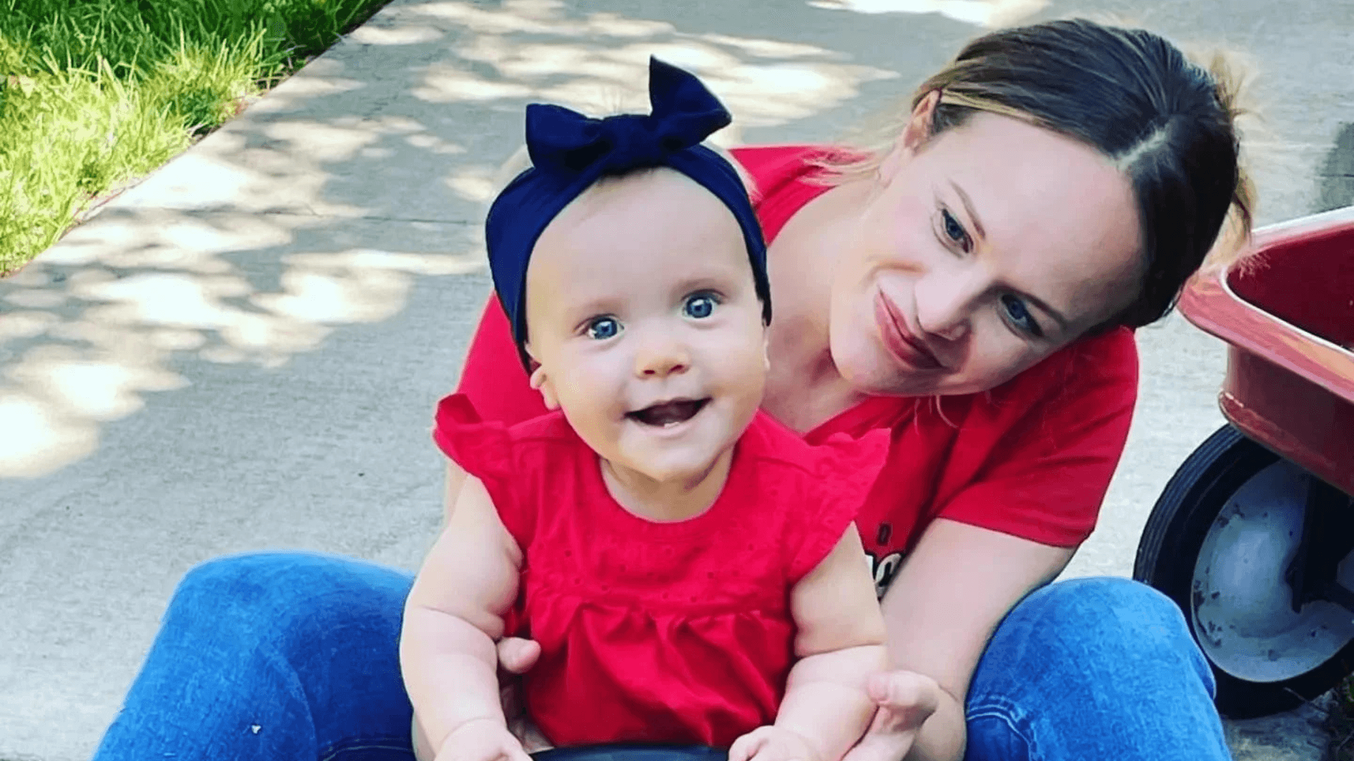 Mother sitting outdoors on a sidewalk holding her smiling baby dressed in red with a matching bow headband, with a red wagon nearby.