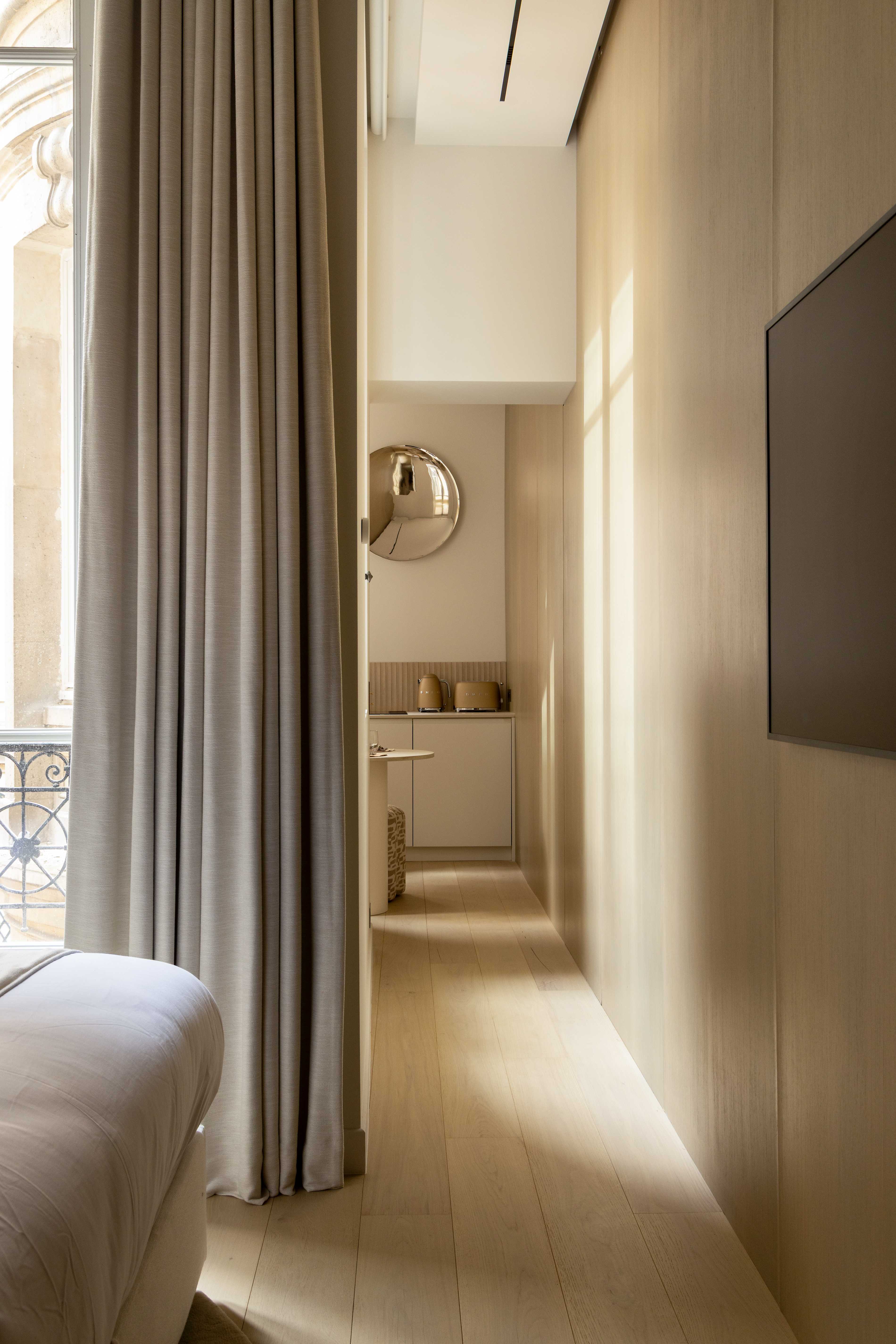 Perspective depuis la fenetre vers la kitchenette au fond avec miroir chrome, rideaux beiges et parquet clair