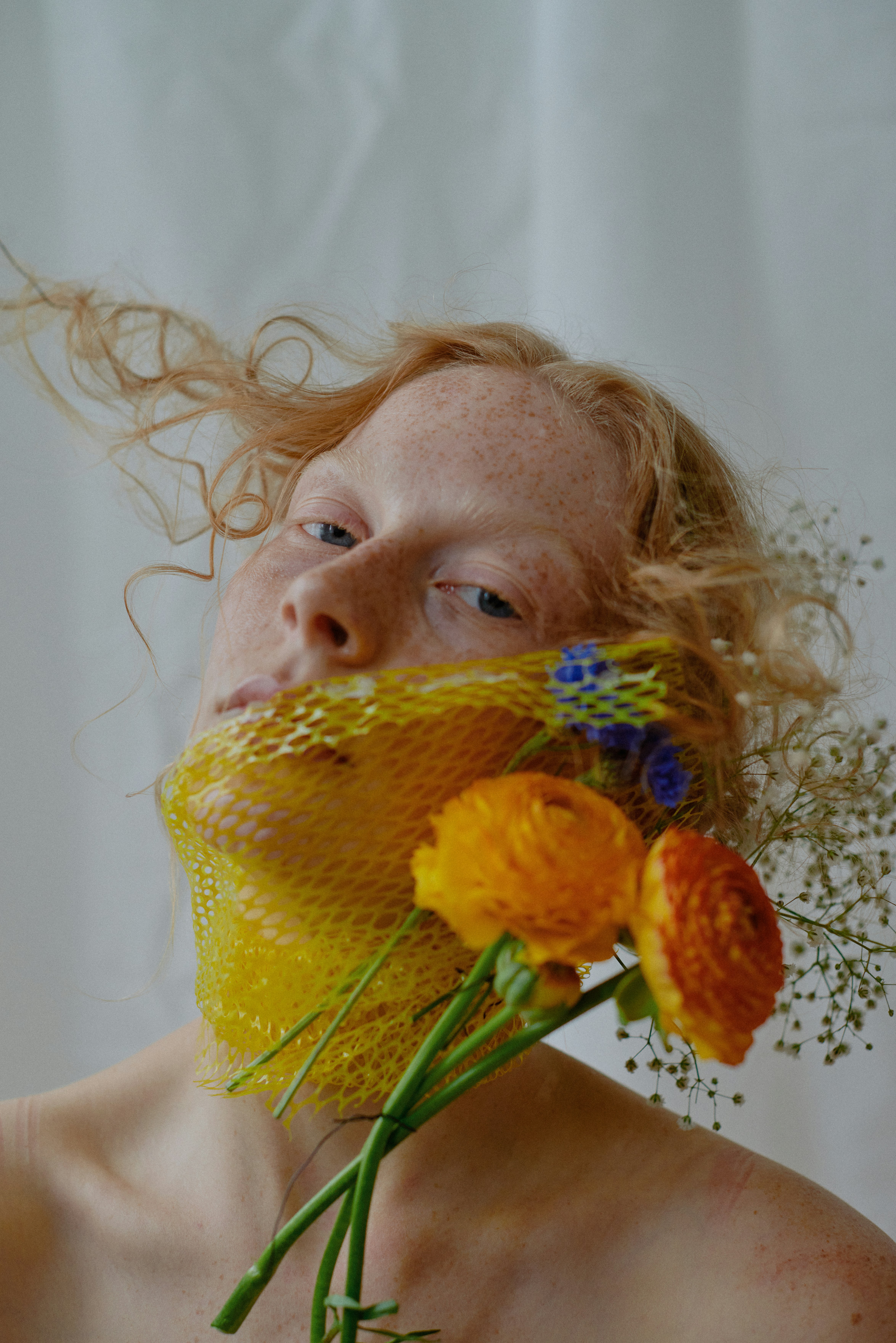 Portrait of a freckled redhead model with yellow mesh and orange ranunculus flowers held against her face, shot against a white backdrop