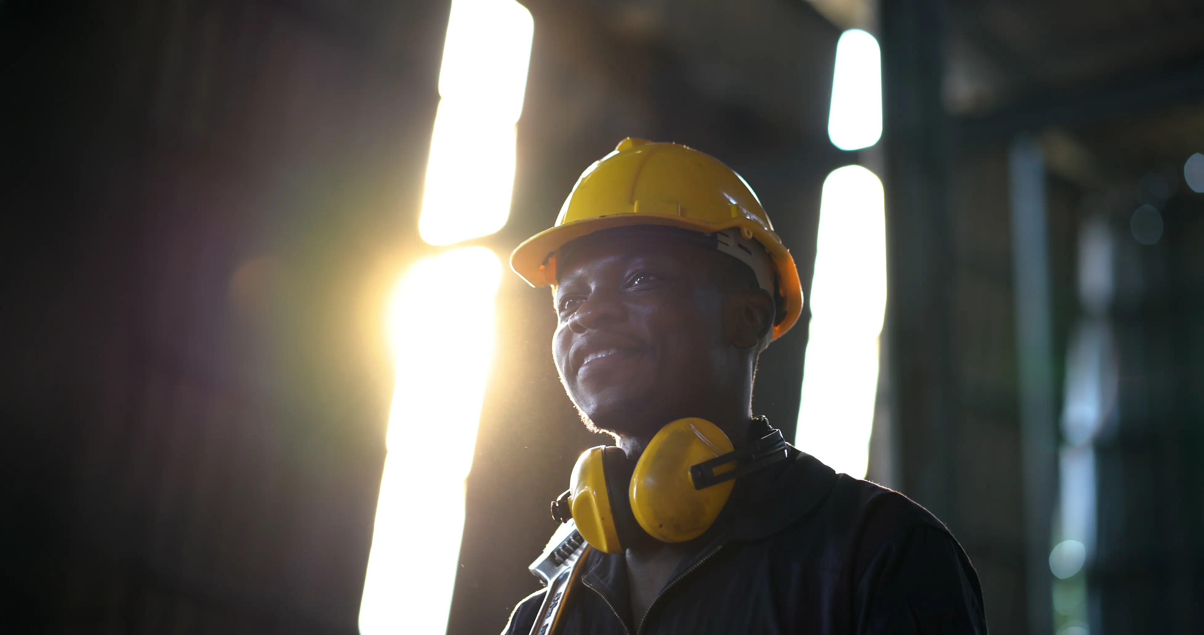 Smiling young man wearing a yellow hard hat and protective headphones stands in a softly lit industrial setting.