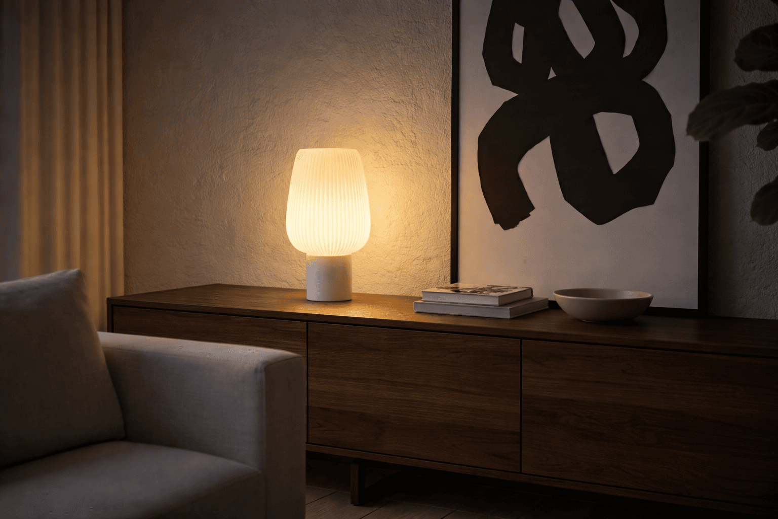 A warm, glowing table lamp with a ribbed white shade sitting on a dark wood sideboard next to abstract wall art.