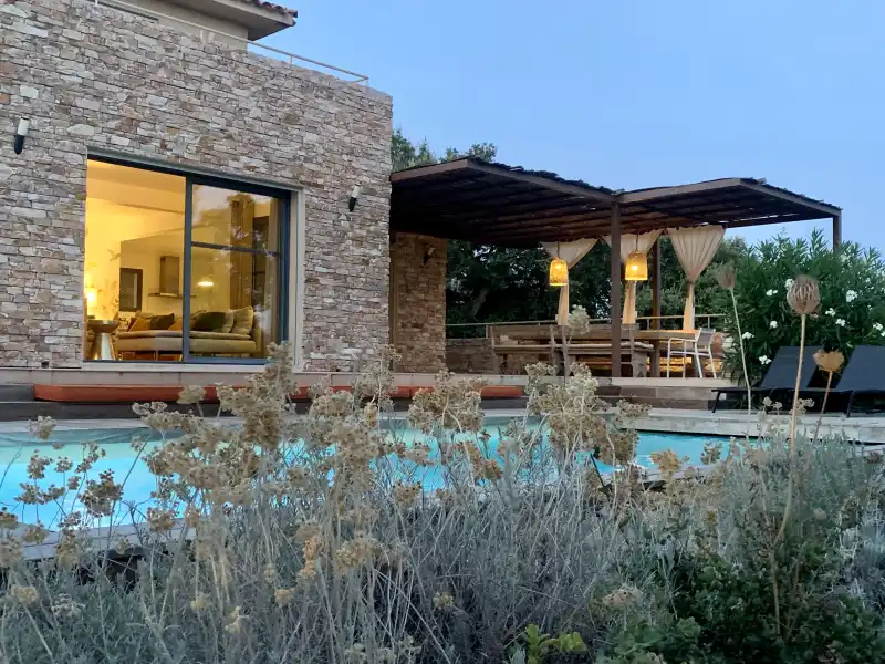 Façade en pierre d'une villa familiale en Corse au crépuscule, piscine illuminée et pergola avec guirlandes lumineuses, projet de décoration intérieure