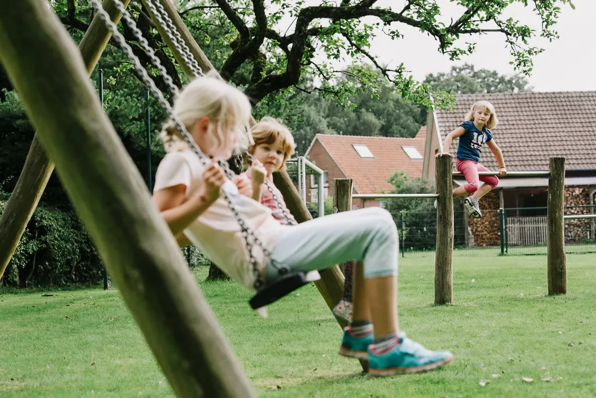 Drei spielende Kinder auf einer Schaukel und einem Spielplatz. Mädchen schwingt, zwei Kinder schauen zu und eines springt.
