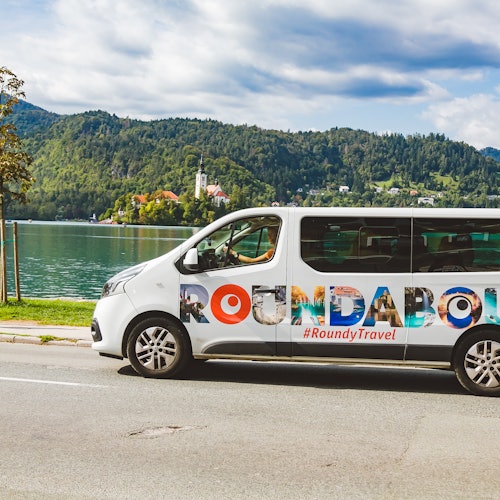 רכב מסחרי לבן עם הכיתוב "ROUNDABOUT" ו-"#RoundyTravel" חונה ליד אגם עם גבעות ועצים ברקע.
