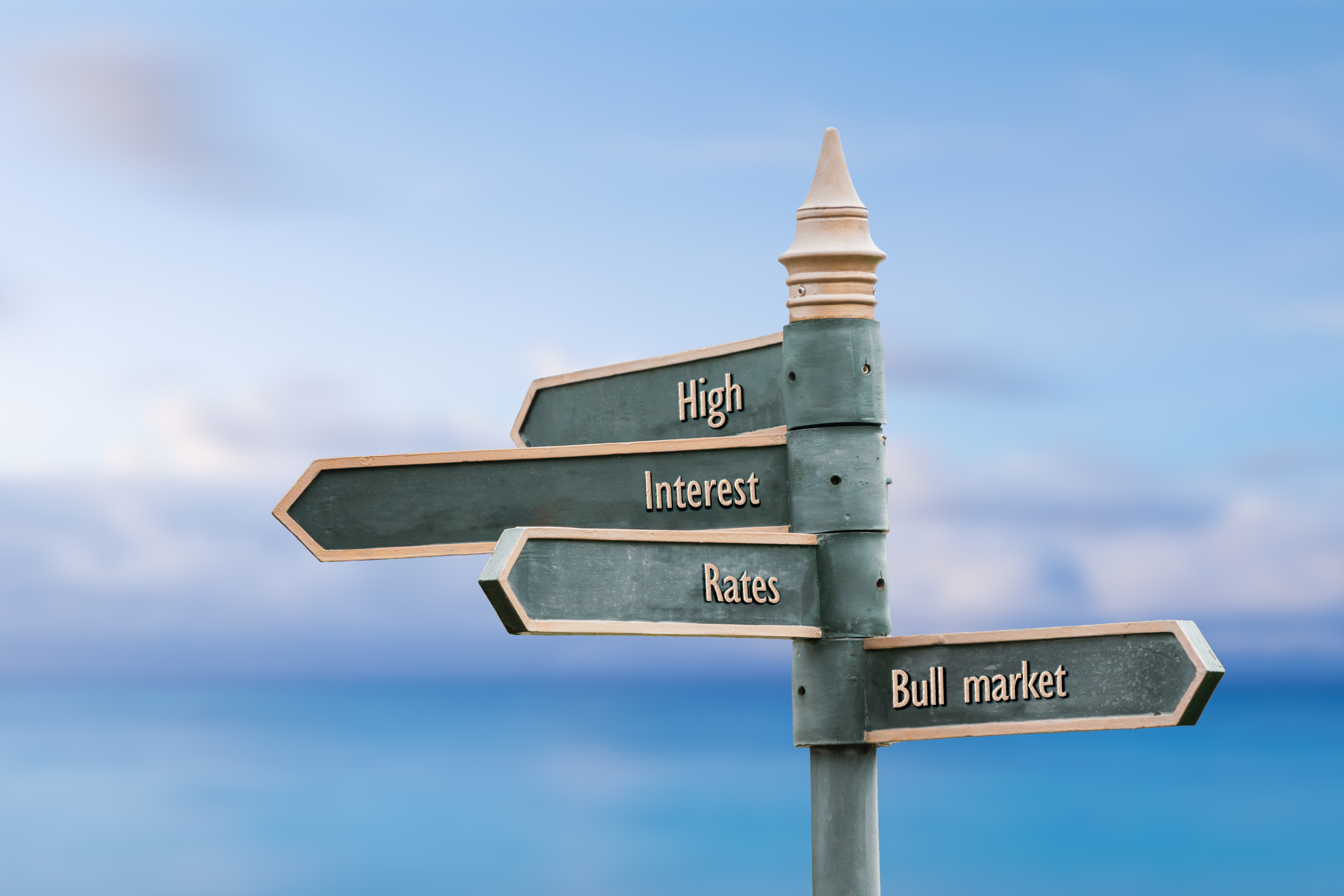 Panneau directionnel vert en acier au bord de la mer, indiquant les mots « high », « interest » et « rates » dans une direction, et « bull market » dans l’autre.
