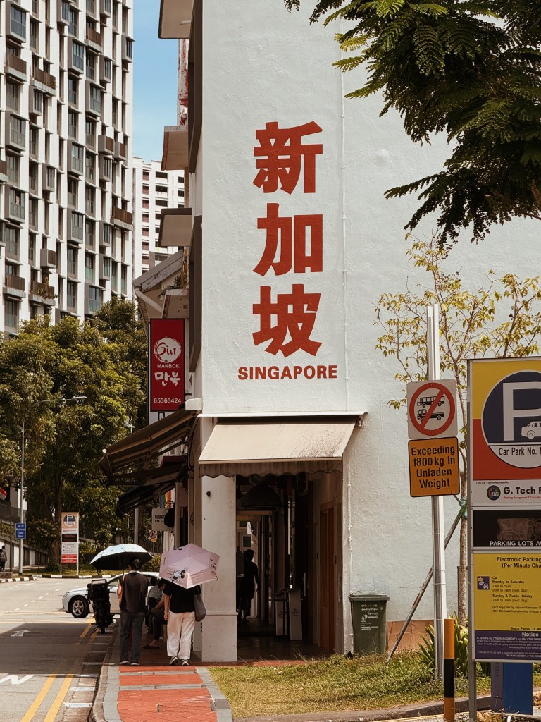 A street view in Singapore featuring a building facade with Chinese characters and 'SINGAPORE' in English, framed by trees and pedestrians holding umbrellas.