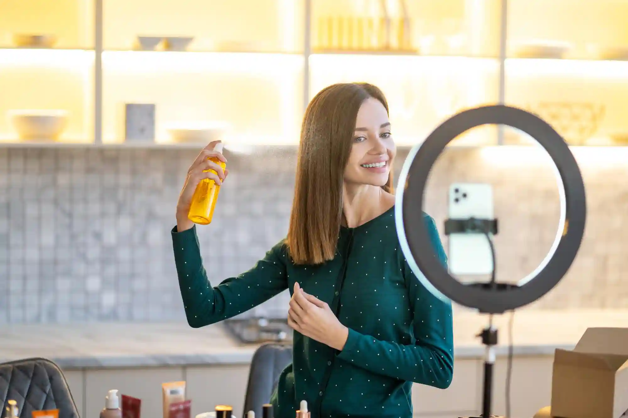 A woman in a kitchen uses a ring light and smartphone to film a beauty product review video.