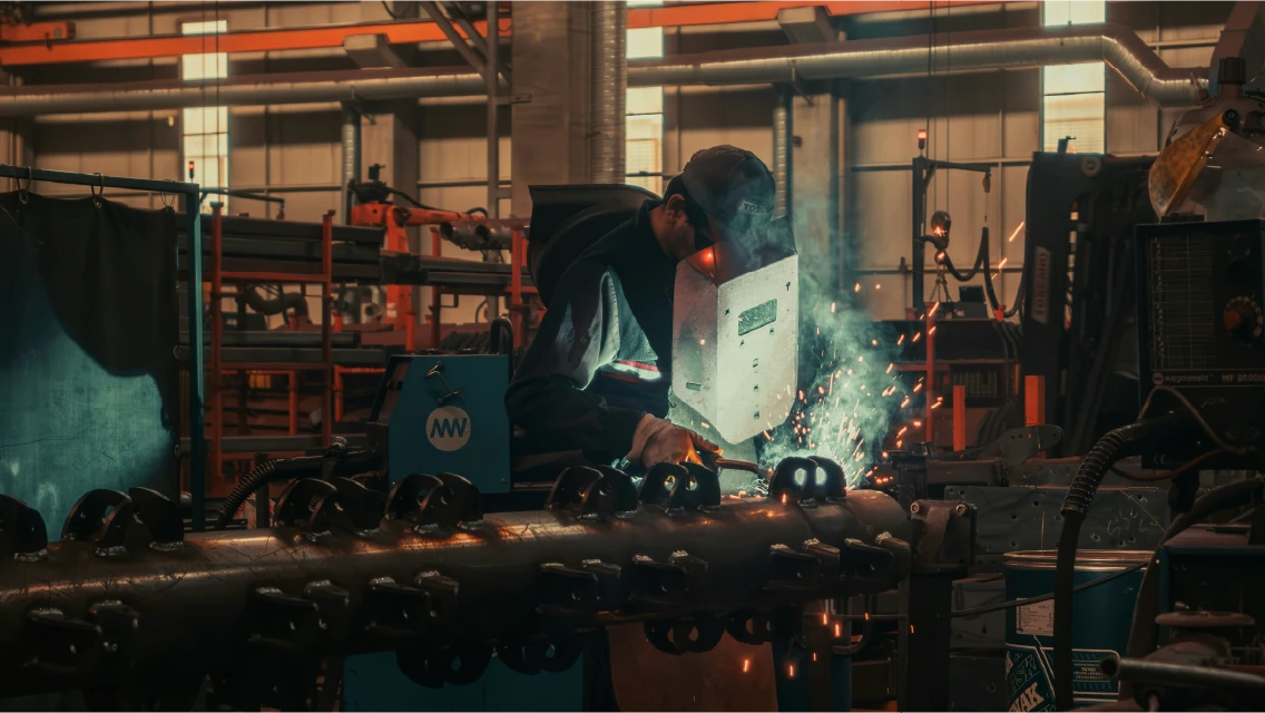 A welder works on heavy machinery in an industrial facility, with sparks flying and welding equipment visible in the background.