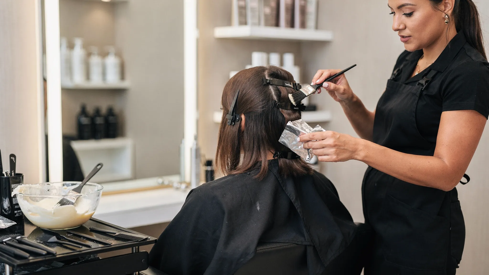 A professional colorist in a modern salon applying lightener to hair using foils and a tint brush, with clean sectioning clips and a bowl of product on a station.