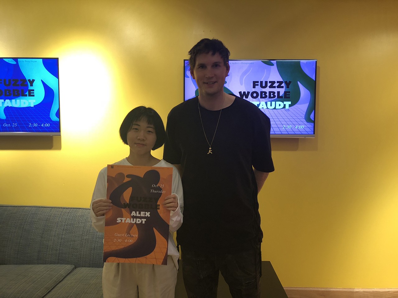 A photo of two people, one holding an orange poster with some figures on it. They both are standing in an exhibition room and behind them on the yellow wall there are some lcd screens with artwork.