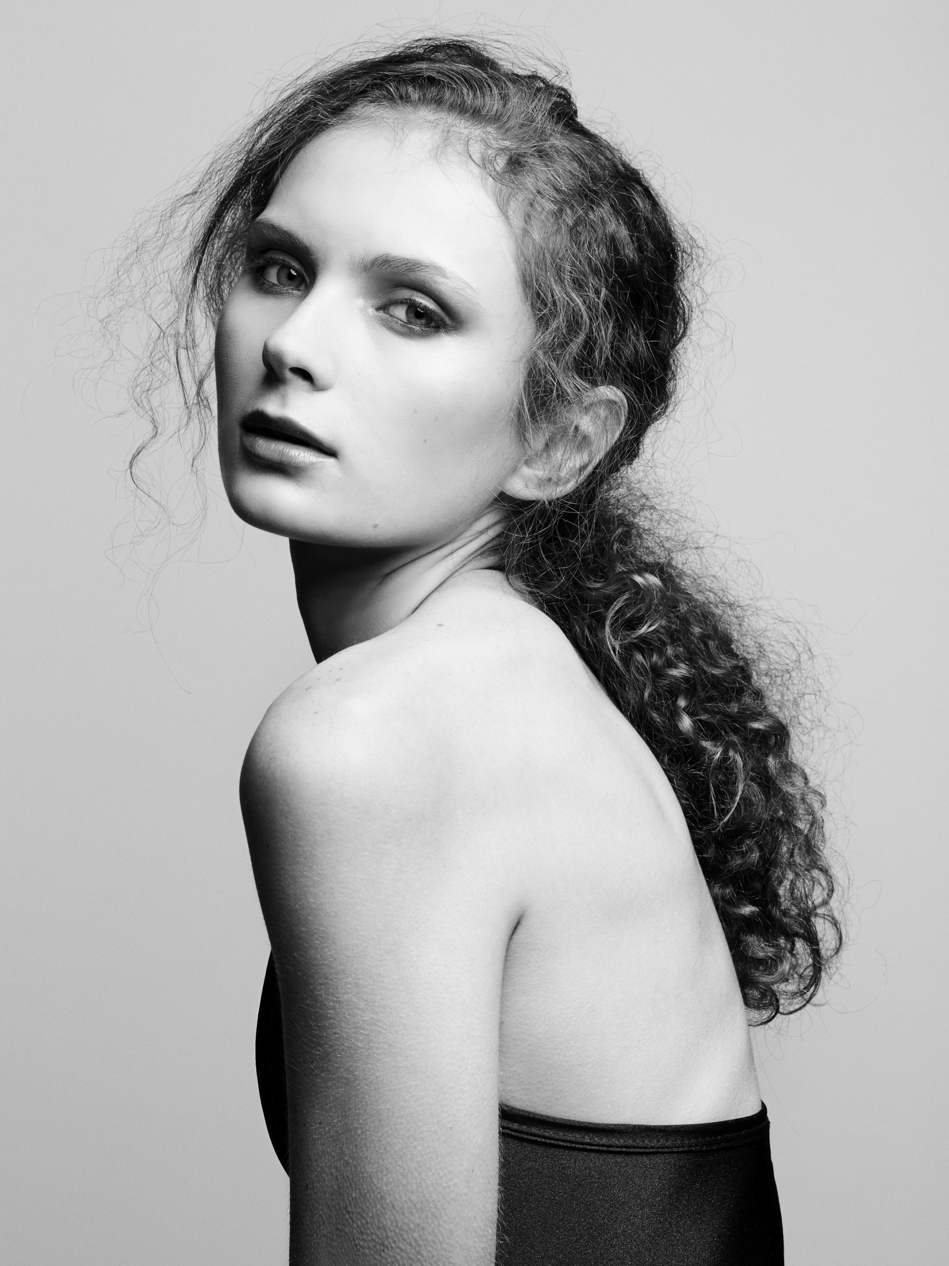 A black and white portrait of a female model with tied-back long curly and bare shoulders looking back toward camera on a minimal light grey background.