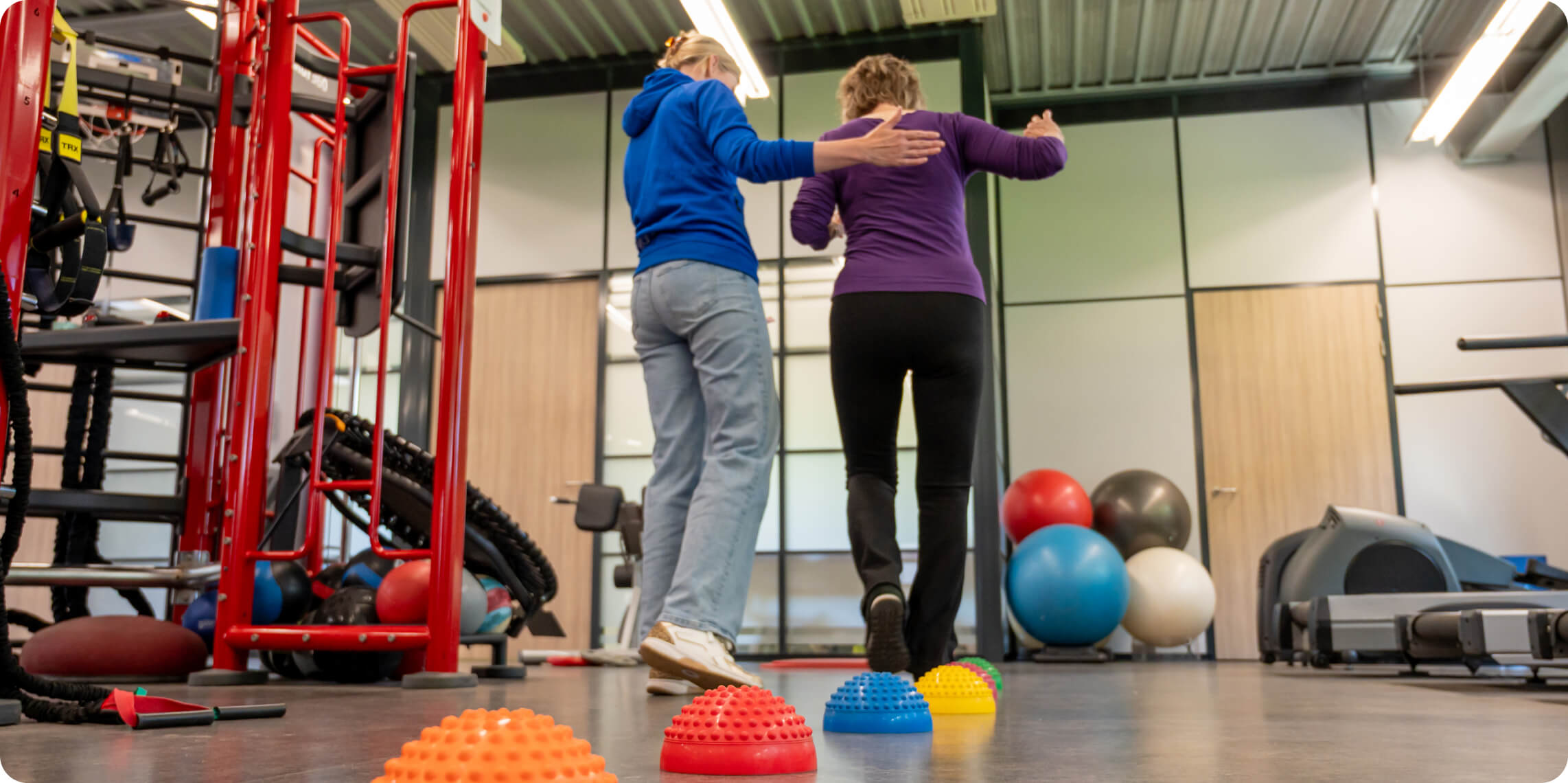 Two people in athletic wear are navigating a rehabilitation gym, focusing on balance exercises using vibrant dome-shaped balance pods on the floor, surrounded by fitness equipment like exercise balls and resistance machines.