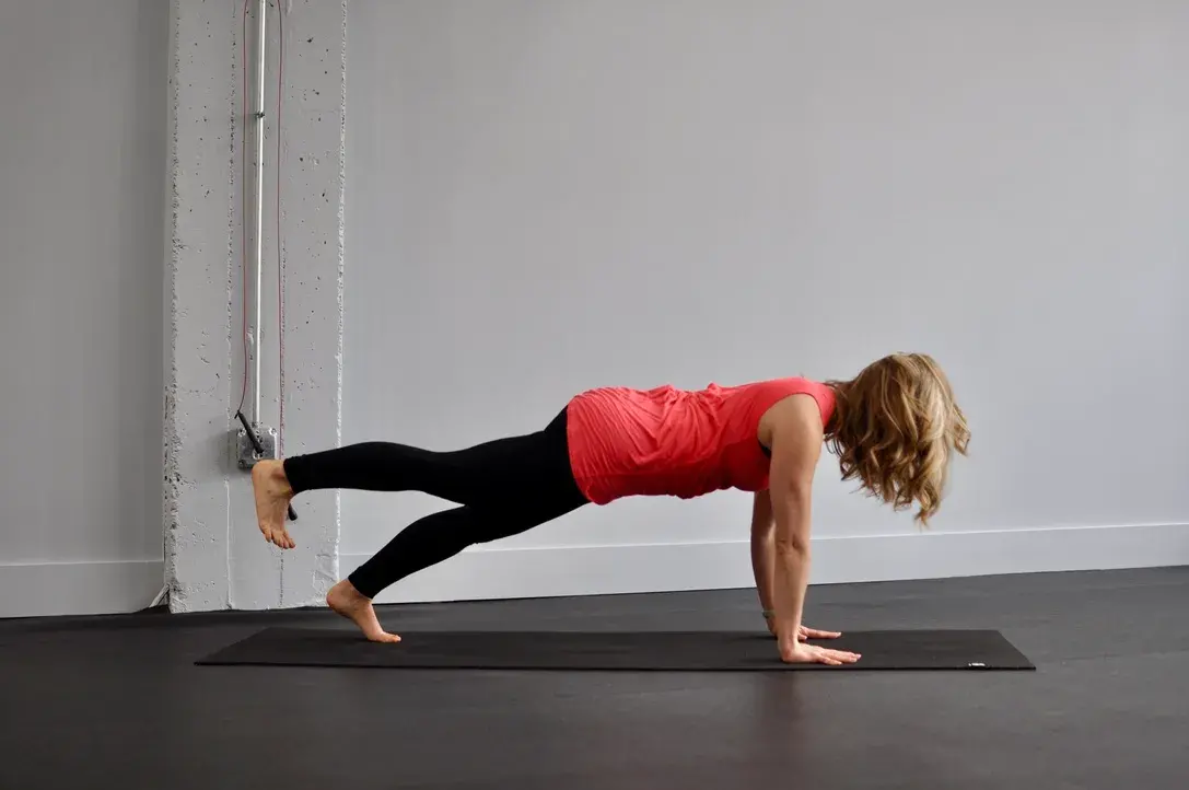 Heather Hausman performs a pilates exercise by balancing in a plank position on one leg, while wearing a red tank top and black leggings, in a minimalistic room with gray walls and a yoga mat.