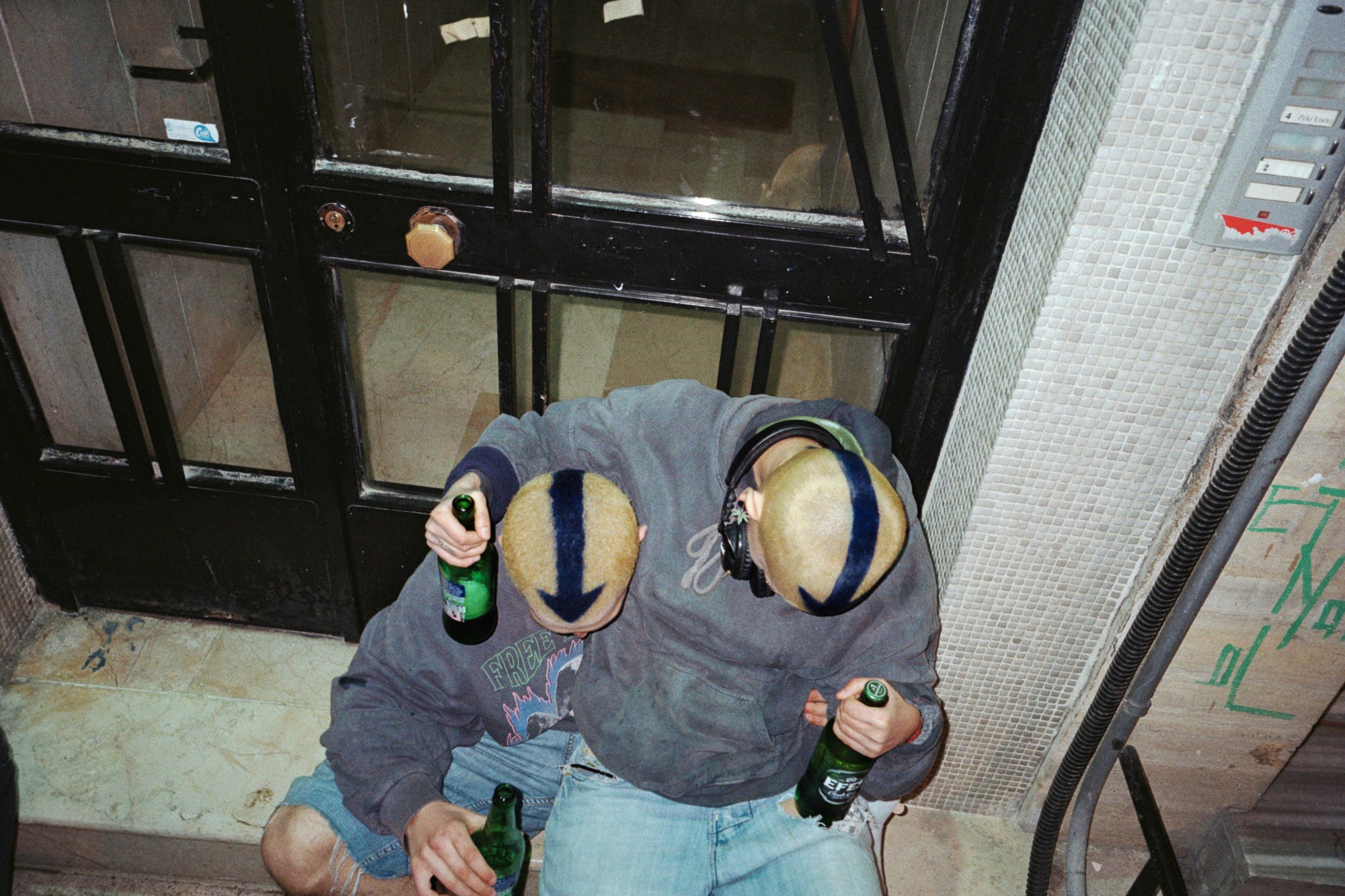 Two individuals with striped haircuts and wearing hoodies sit outside a building entrance, each holding a green bottle, conveying a relaxed urban setting.