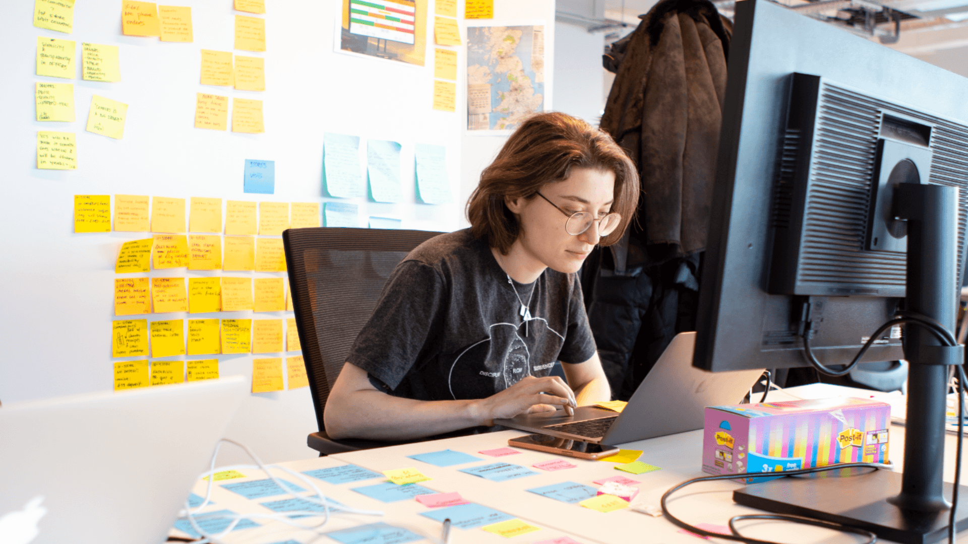 A woman sits at her desk, working on a computer, infront of a wall covered with sticky notes