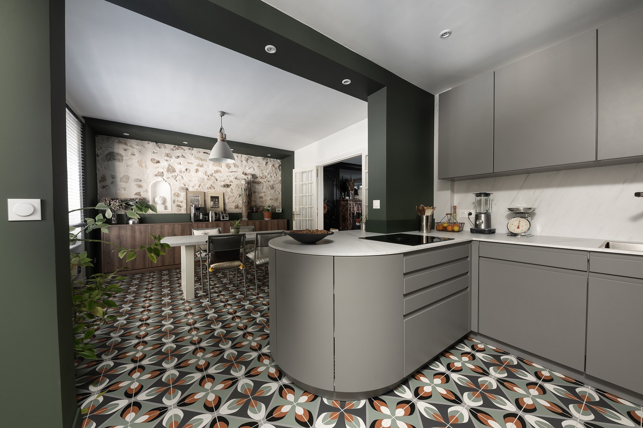 Cuisine moderne avec des armoires grises, un comptoir en marbre, un sol en carrelage à motifs et des murs vert olive. Agencement ouvert avec un coin repas, des lampes suspendues, des plantes et un mur d'accentuation en pierre à l'arrière-plan.