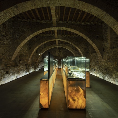 A dimly lit stone hall with arched ceilings featuring illuminated display cases with artifacts.
