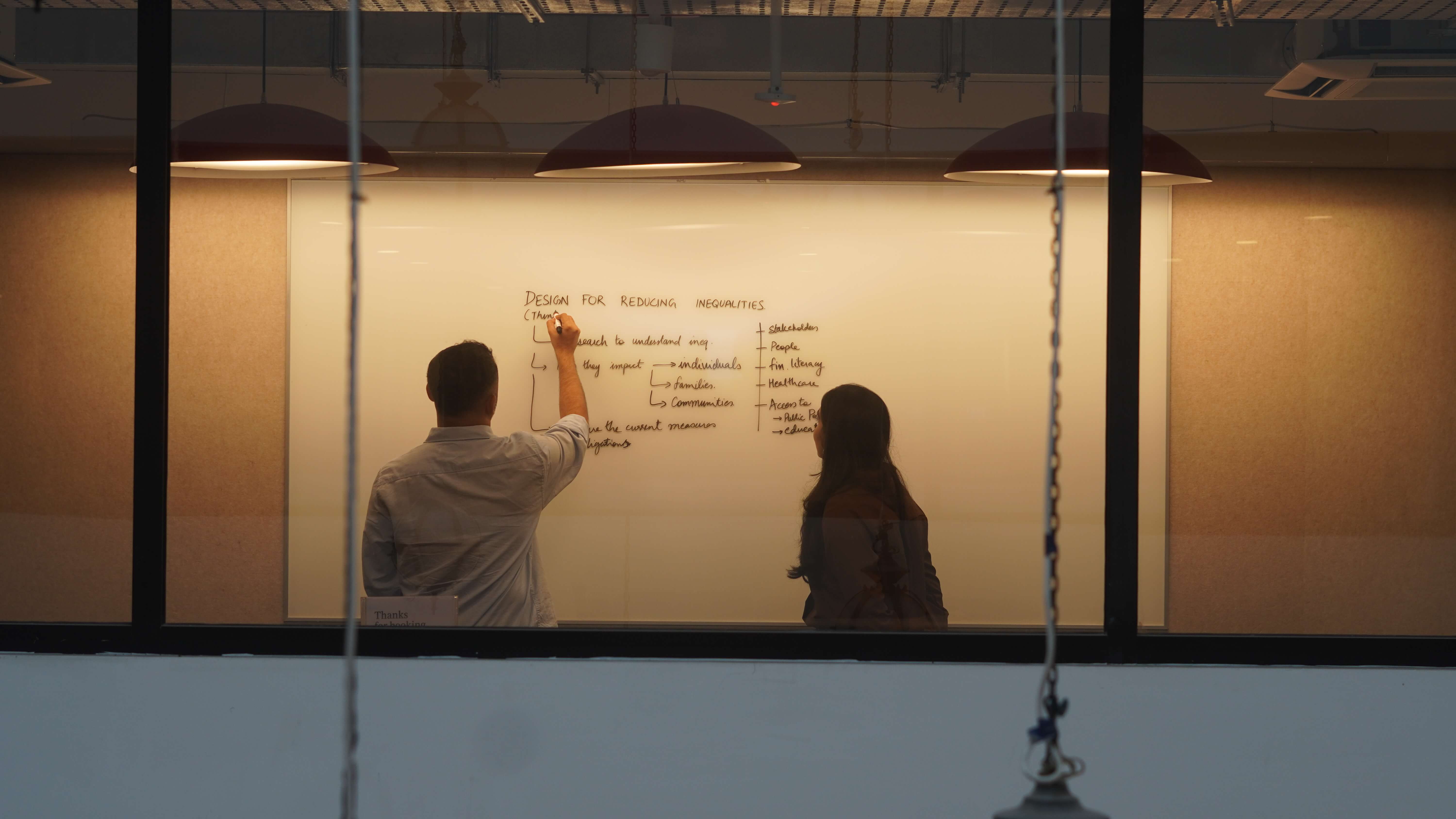 A man stands while two people sit at a table in an office setting, smiling and engaged in conversation. The mood is friendly and collaborative.