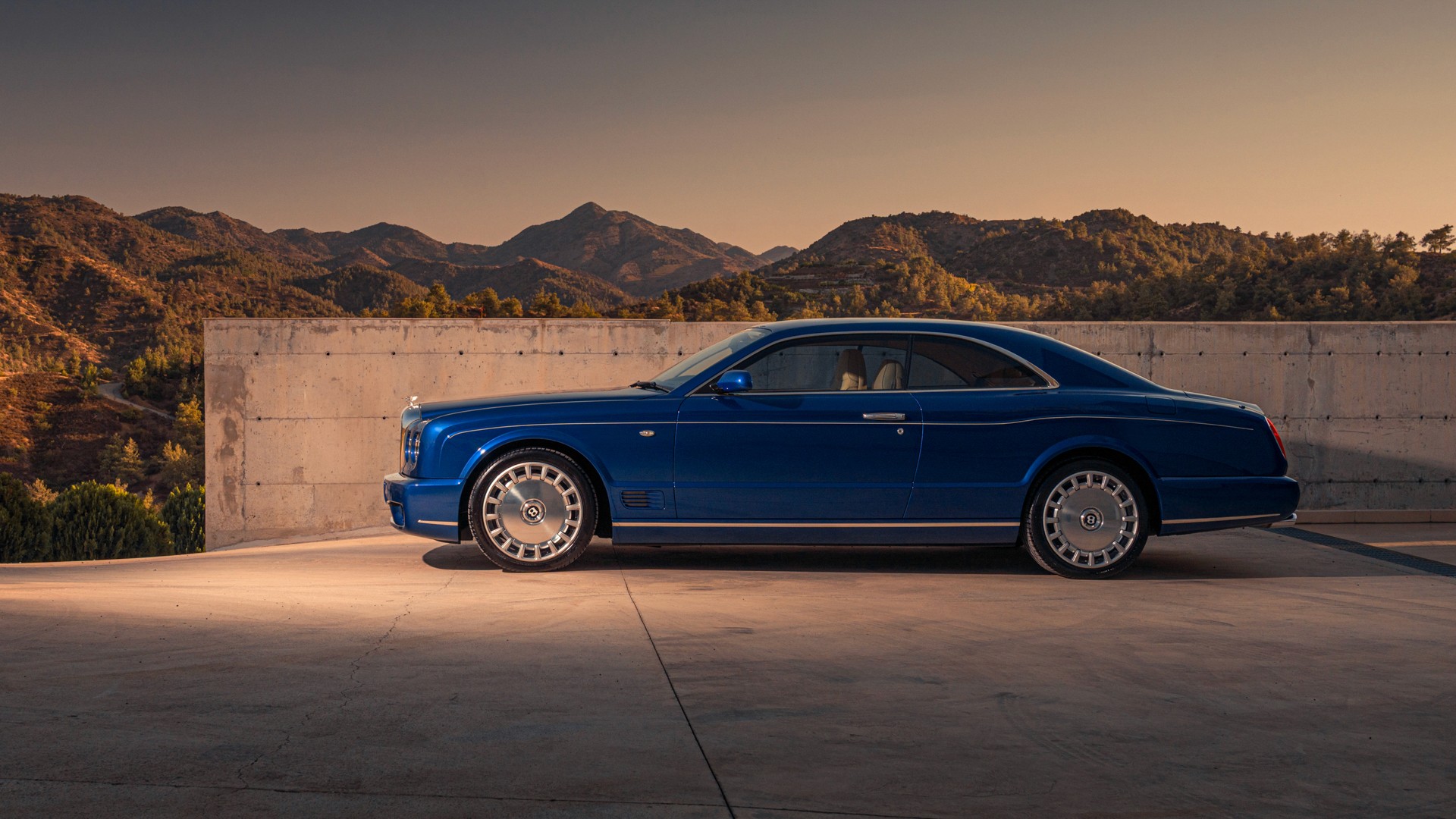 Bentley Brooklands coupé in Moroccan Blue, shot in side profile on a concrete platform in the foothills of Troodos, Cyprus, mountains and golden-hour light behind, polished multi-spoke wheels catching the low sun.
