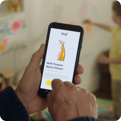 A person holding a smartphone in their hands, displaying a delivery app showcasing a multi-purpose spray cleaner, with a blurred background of children’s drawings on a wall.