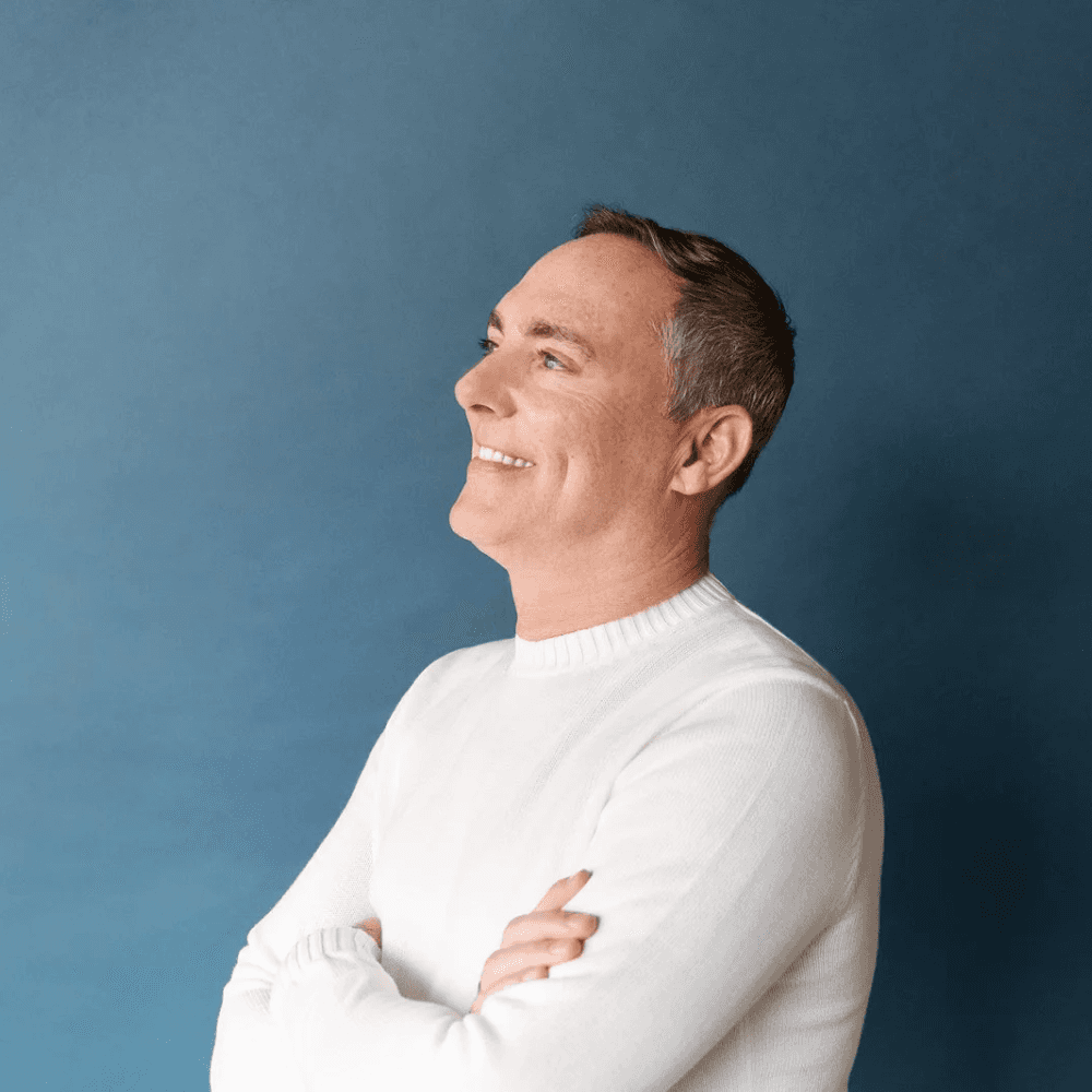A man in a white sweater stands against a blue background, smiling with arms crossed. He gazes upward, conveying a positive and confident mood.