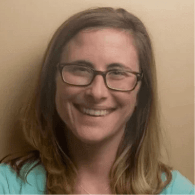 Alt tag: Portrait of Krista Lehde smiling at the camera, wearing glasses and a teal top against a neutral background.