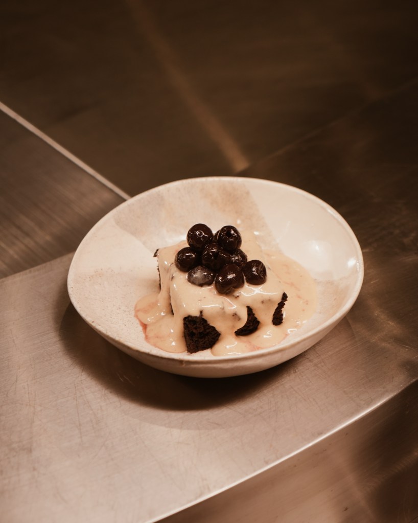 A chocolate brownie topped with cream sauce and black cherries, served in a white bowl against a metallic surface.