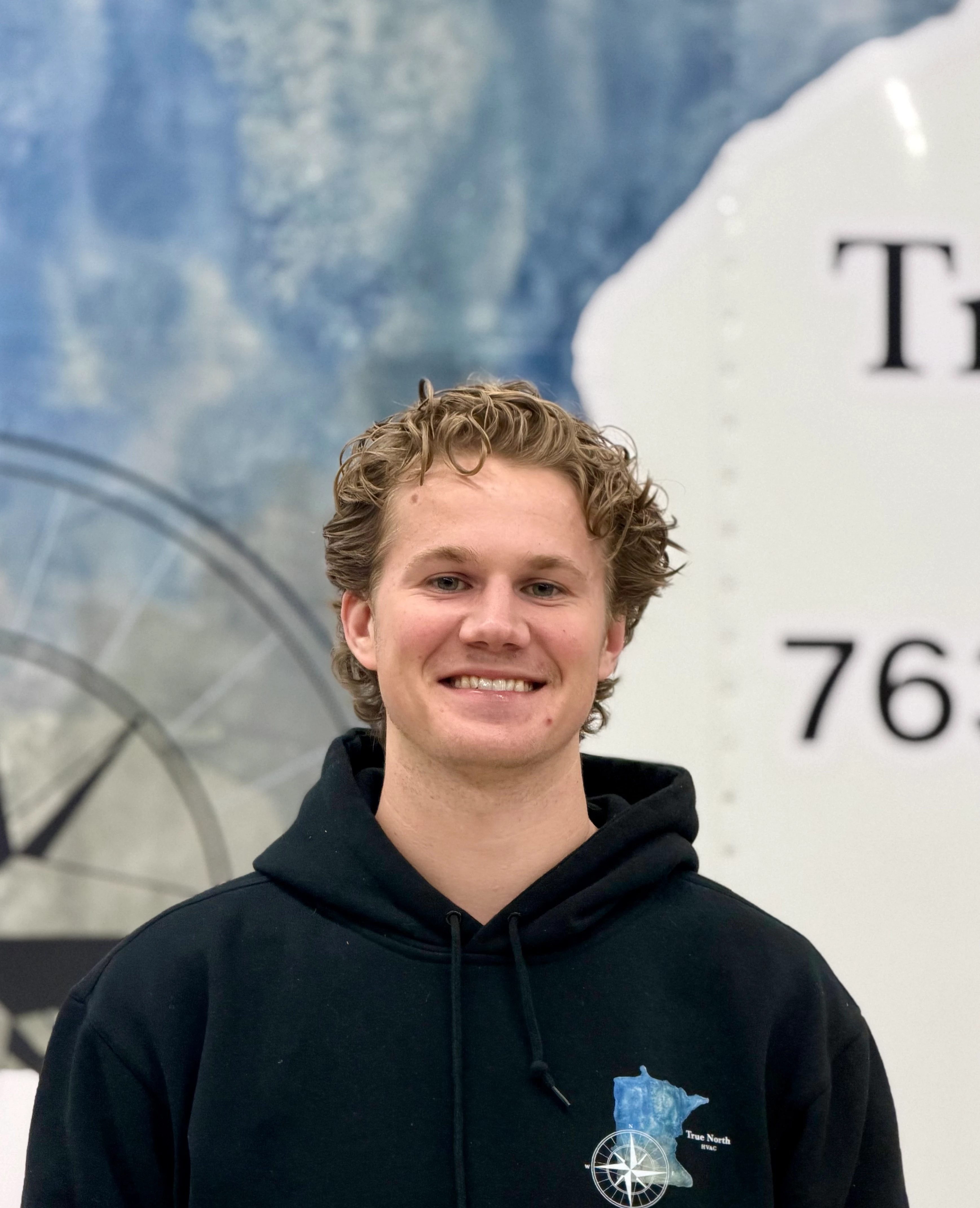 A young woman with long hair smiles while standing against a green backdrop, wearing a casual dark sweater.