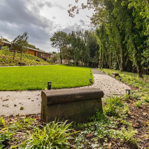 A curved path bordered by grass and trees, with small lights along the edge and a bench in a park setting.