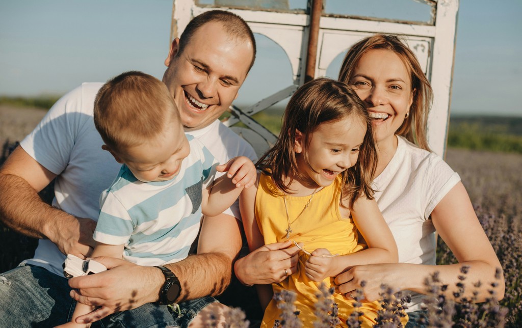 A joyful family of four enjoys a sunny day outdoors, with the parents and their two young children laughing and embracing each other in a lavender field, creating a heartwarming and candid moment.