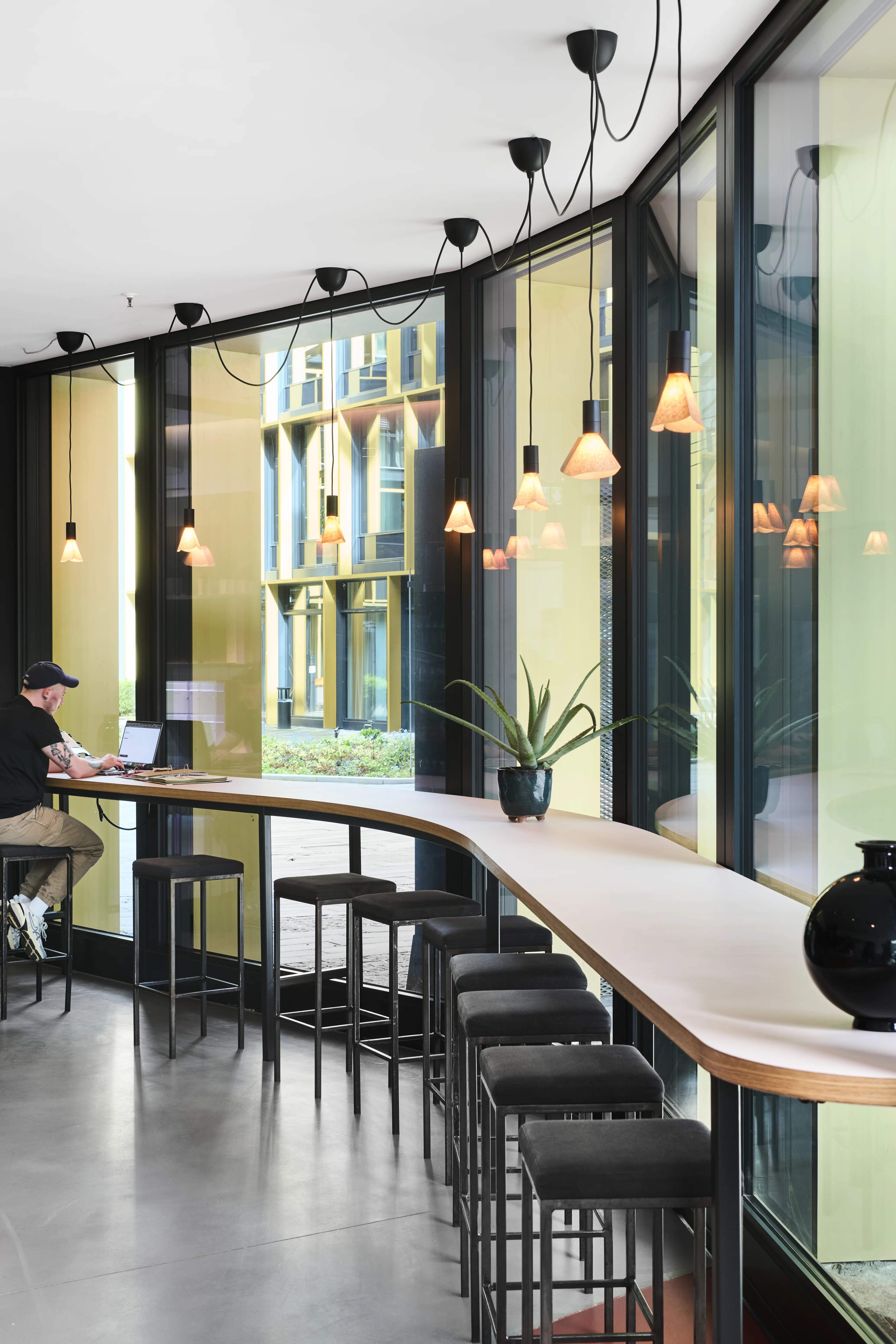 Curved window counter at CAFÉ CAMILLA with a white high table, black stools and black cone pendant lights in front of large arched windows.
