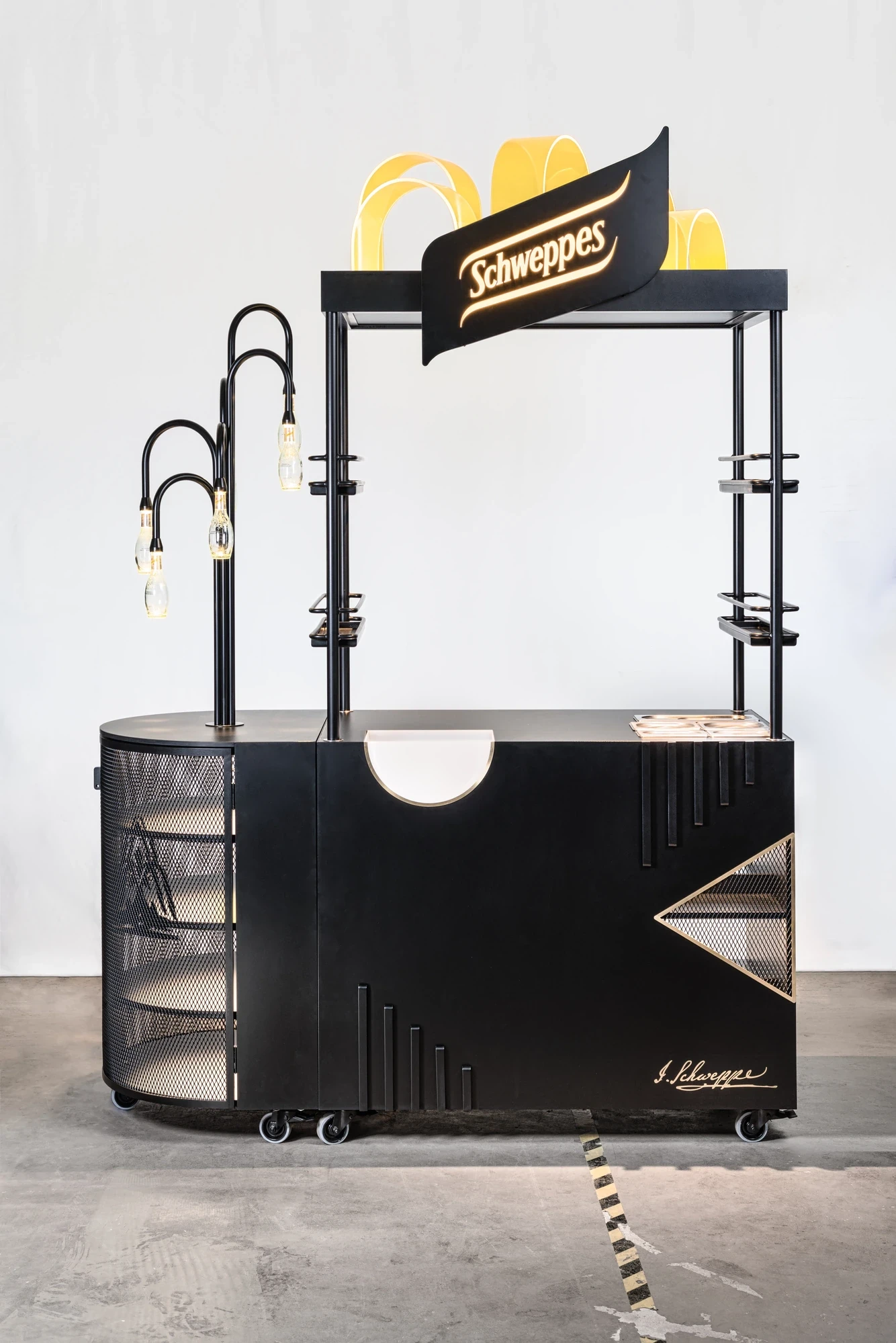 Branded Schweppes mobile bar trolley in matte black with gold trim, flag-shaped logo header, integrated glass shelving and gooseneck lamps.