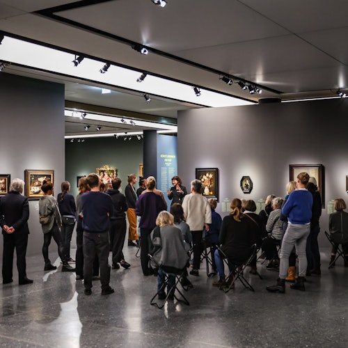 A group of people is listening to a guide in an art gallery, surrounded by framed paintings on dark gray walls.