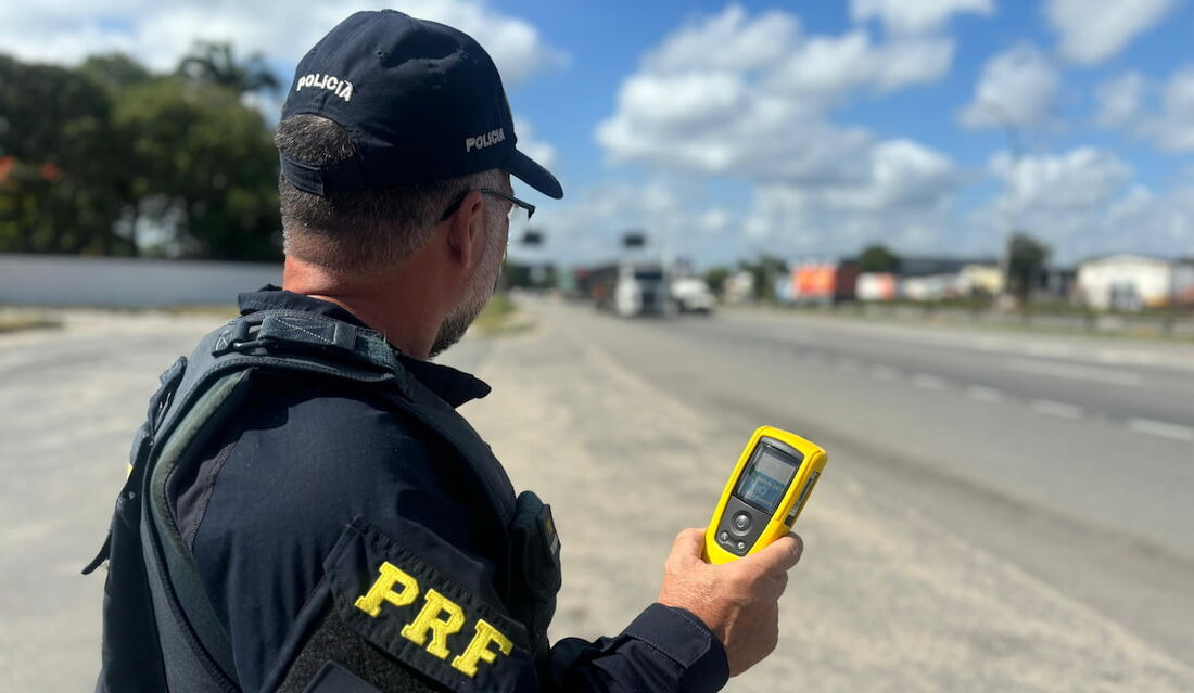 Policial da Polícia Rodoviária Federal (PRF) segurando etilômetro Alcolizer LE5 em operação de fiscalização viária.