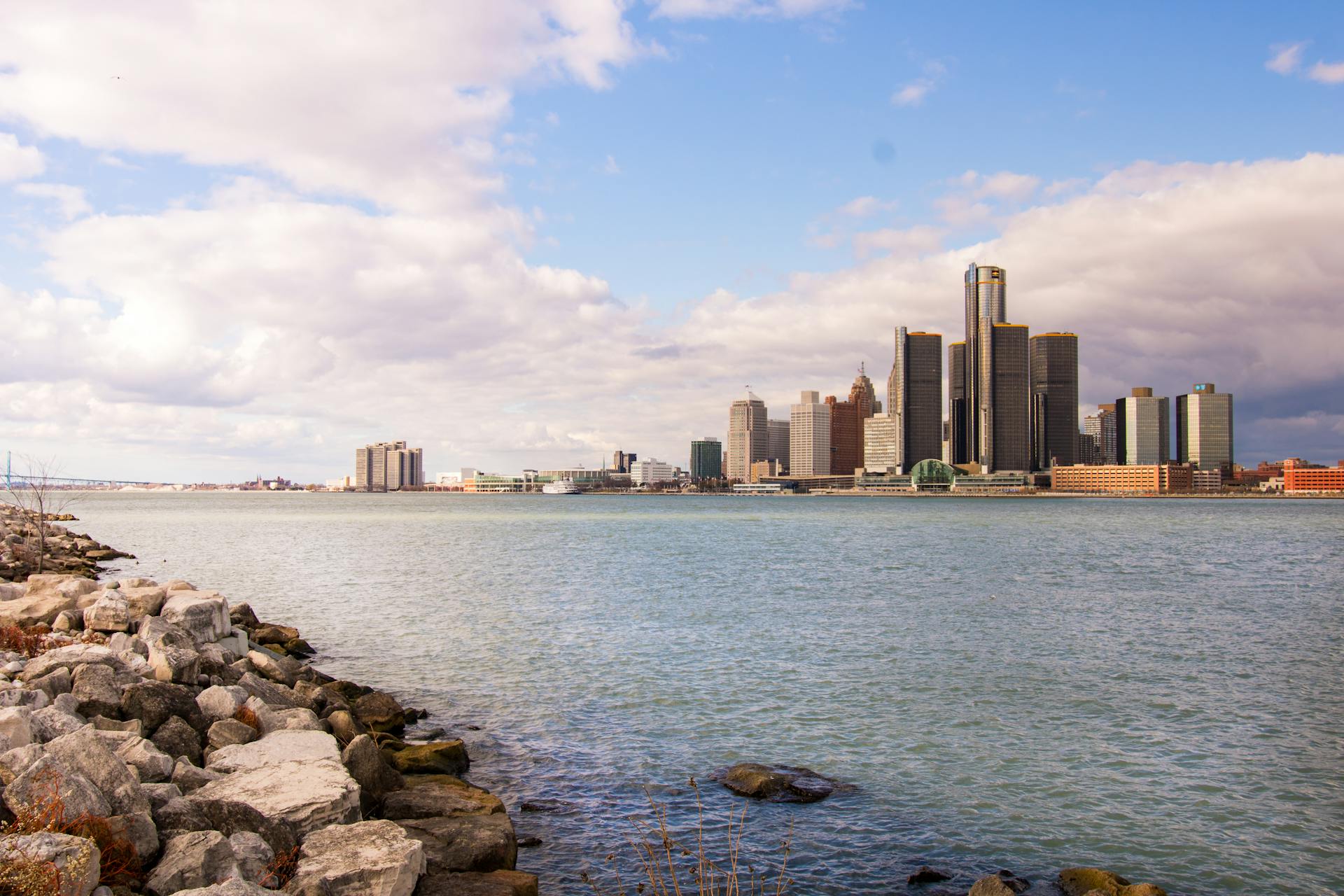 City skyline view with modern buildings, calm water in the foreground, and a cloudy sky above.