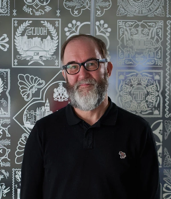 A head‑and‑shoulders portrait of a person wearing a black polo shirt with a small embroidered logo on the chest, standing against a decorative wall featuring patterned illustrations and ornamental designs in muted tones.