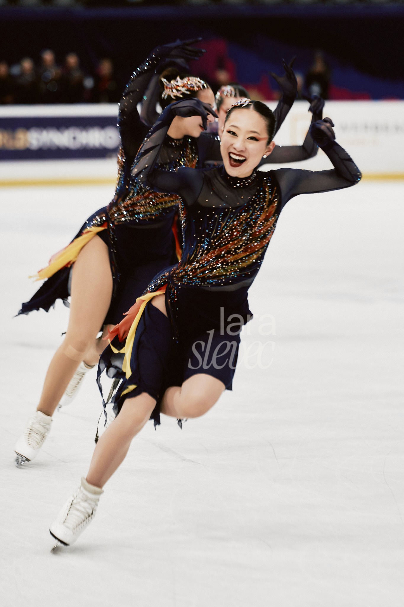 Drei Synchroneiskunstläuferinnen, die auf einem Bein eine Wendung durchführen. Die Vordere Läuferin schaut lächelnd in die Kamera, Hände über dem Kopf. Team Les Supremes. ISU Synchronized Skating Championships 2026 in Salzburg. Fotografiert von Lara Slevec.