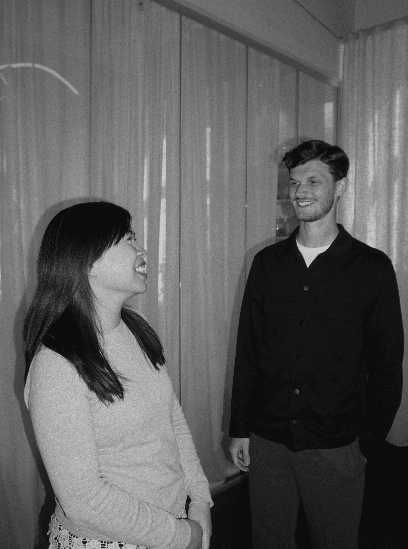 Two people standing indoors, smiling and talking to each other in a casual setting.