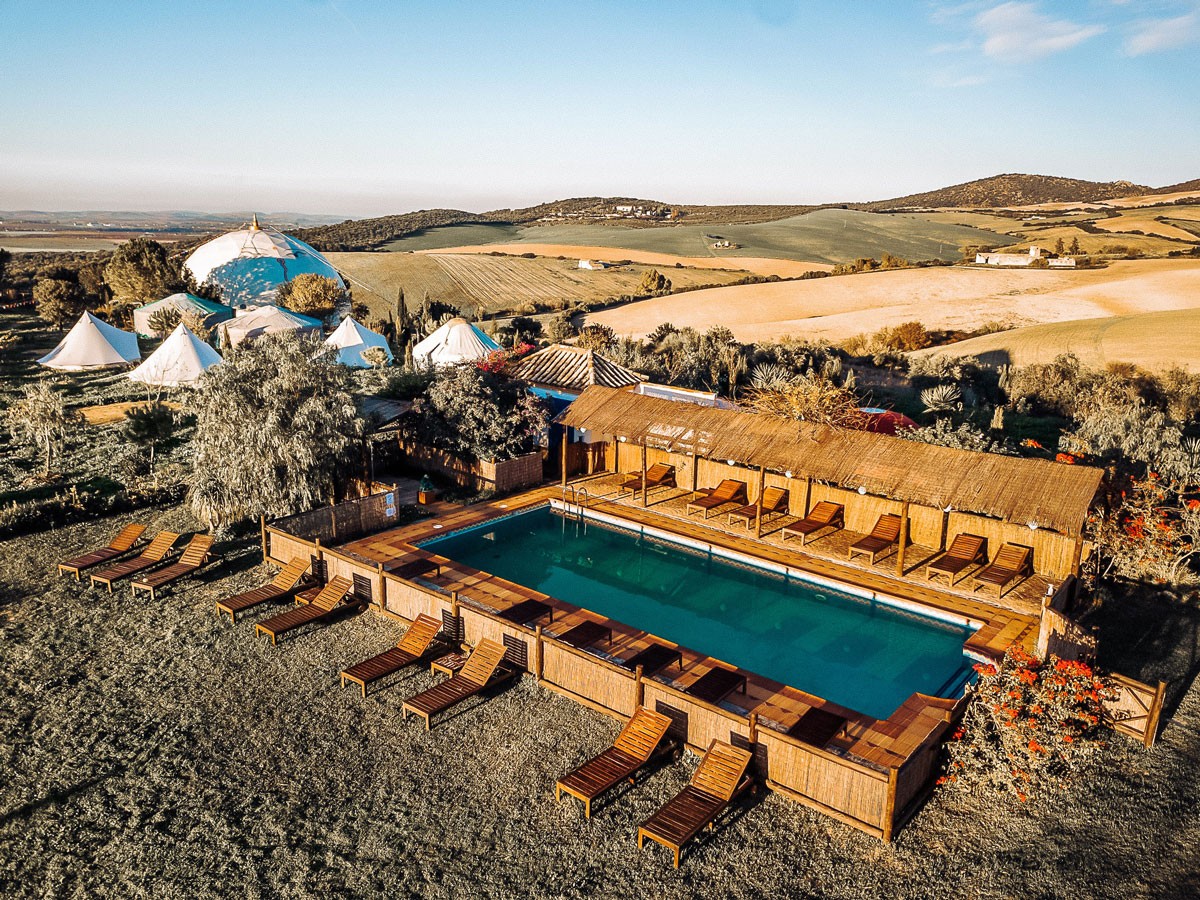 Aerial view of a serene landscape featuring a rectangular pool surrounded by wooden loungers, lush trees, white tents, and rolling hills at sunset.