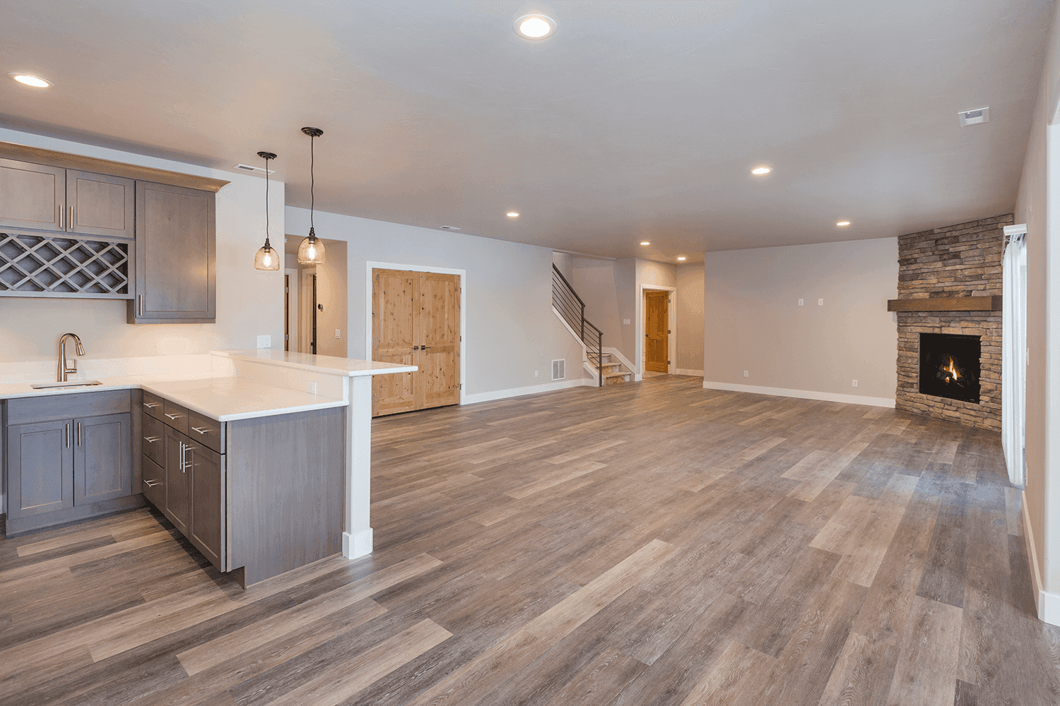 A spacious basement featuring a gray kitchenette with white countertops and pendant lighting, adjacent to a living area with wood-look flooring. A stone fireplace with a wooden mantel and rustic double doors enhance the open, airy design.