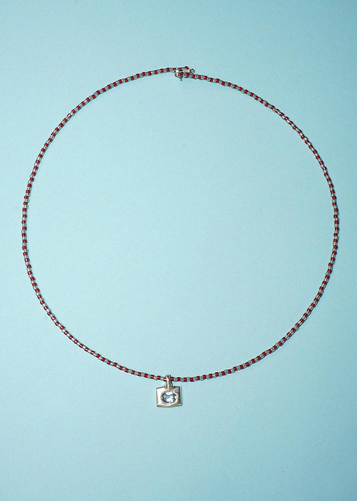 A delicate beaded necklace with alternating red and dark brown beads, featuring a small silver square pendant with a clear central stone, displayed on a light blue background.