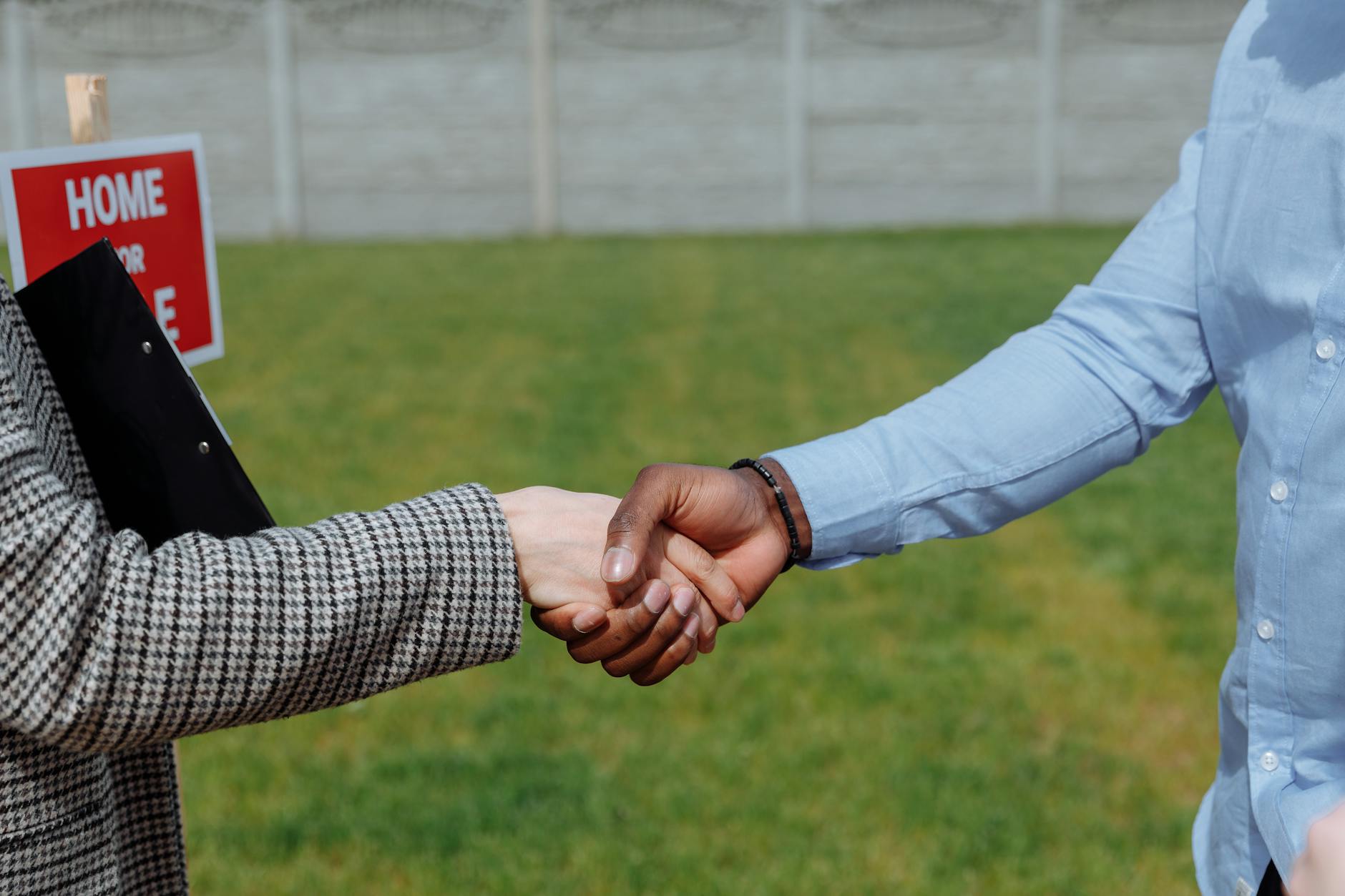 Two people shaking hands outdoors beside a red Home For Sale sign, one holding a clipboard