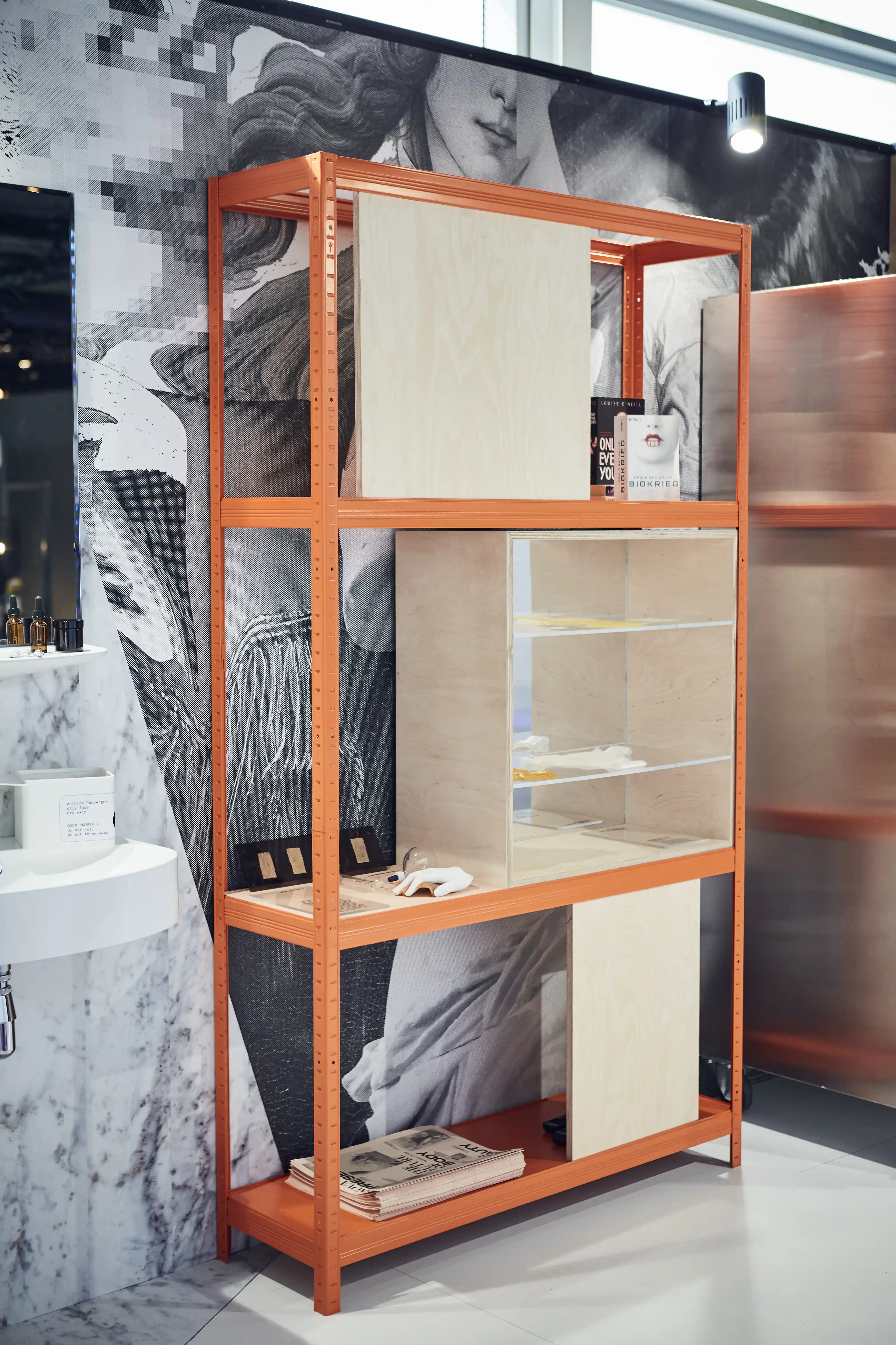 Orange display rack with plywood panels and glass vitrine in front of a pixelated classical-art wall at the Cosnova exhibition.
