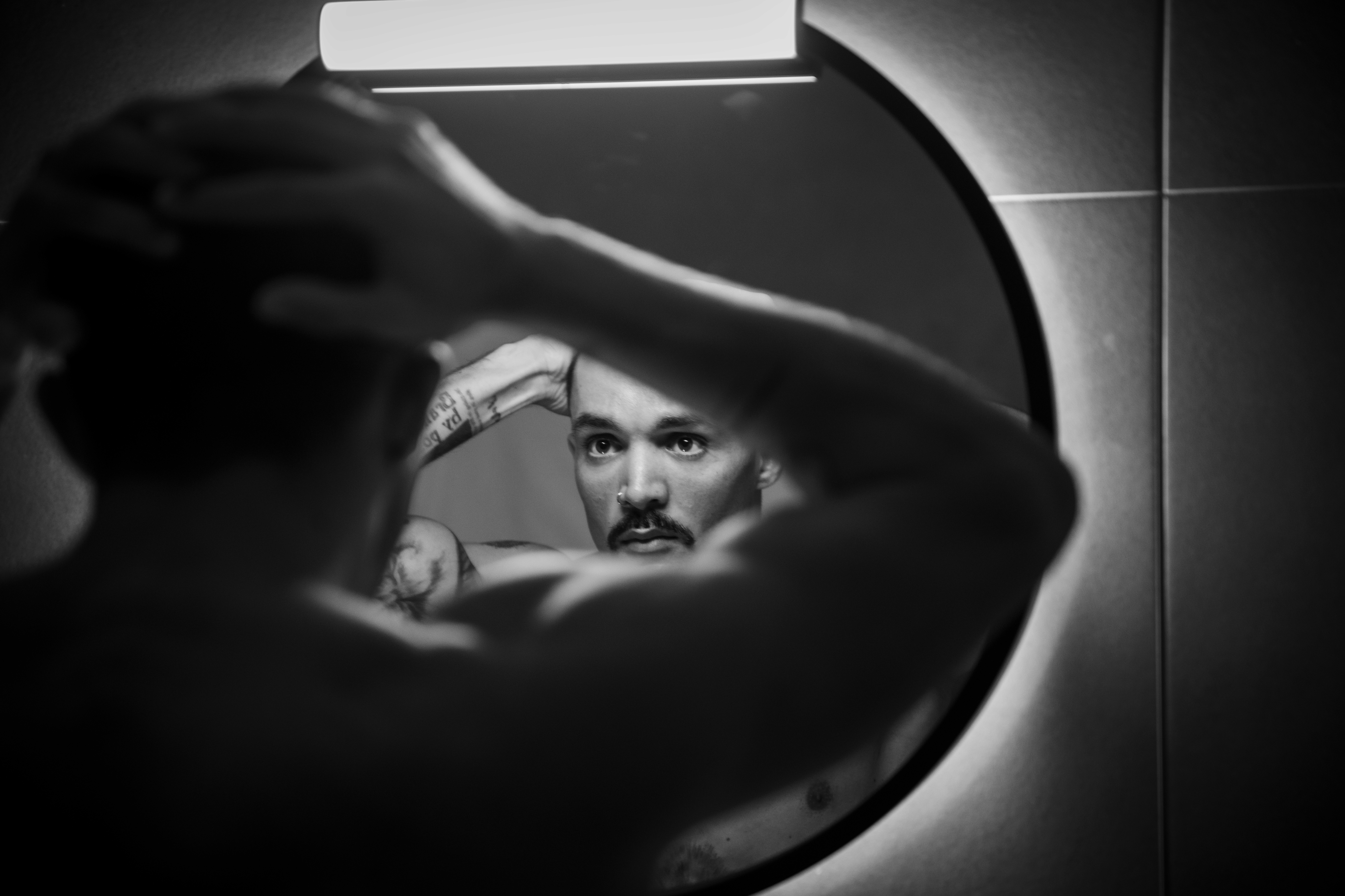 Black-and-white photograph of a man looking into a circular, illuminated mirror while adjusting his hair