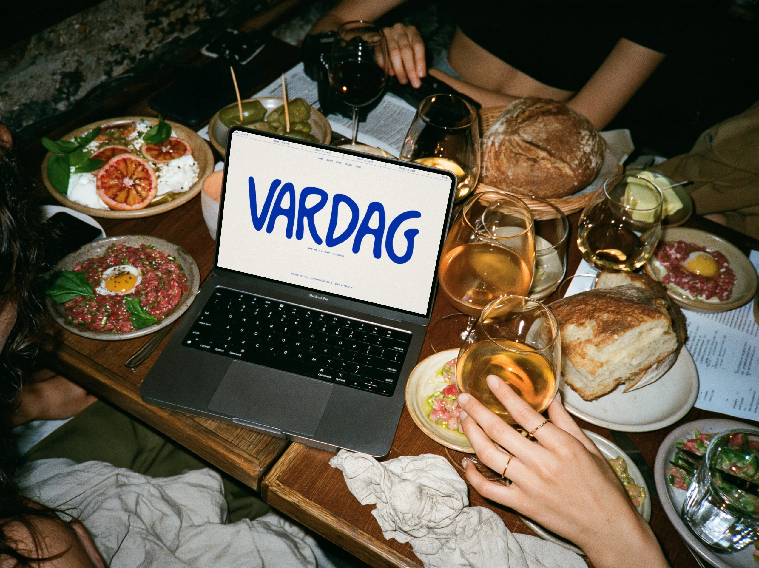 A MacBook on a crowded wine bar table showing the Vardag website, surrounded by small plates, bread and glasses of orange wine.
