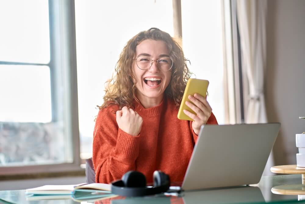 Excited woman holding phone in front of laptop